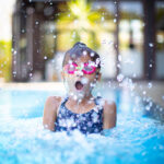A young girl with pink googles on surfacing in an outdoor pool and taking a big breath.