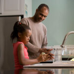 A father and daughter at the kitchen sink; daughter is washing her hands with a bar of soap, father is smiling