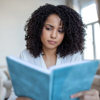 A woman with dark curly hair and wearing a white shirt reading a blue book