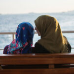 A mother and daughter, both wearing hijabs, seated on a bench and talking
